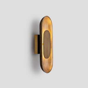 Modern oval LED wall sconce in brushed brass on a deep blue entryway wall next to a round mirror and potted plant.