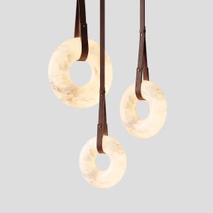 Alt Text: Three illuminated alabaster ring pendant lights suspended by brown leather straps with brass hardware against a light grey background.